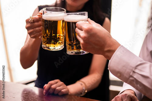 A close-up shot of a man and woman toasting with beer mugs in a warmly lit bar. The image focuses on their hands and glasses, emphasizing the golden color of the beer and the festive atmosphere.