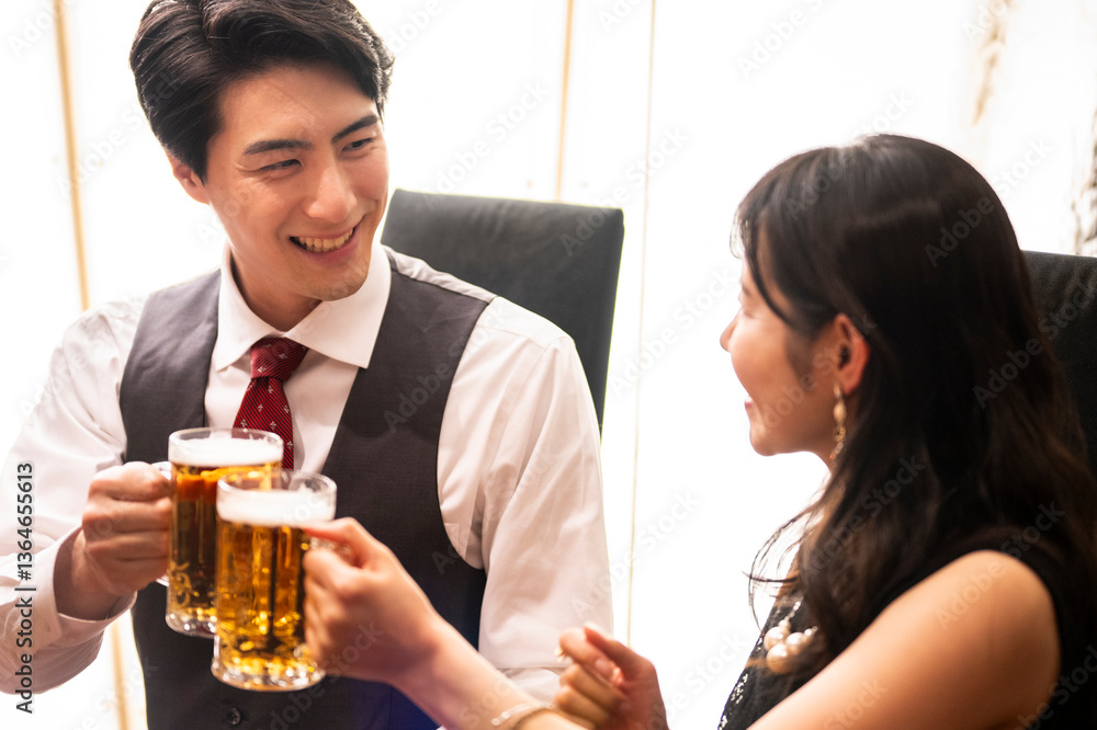 A smiling man and woman enjoy a moment of celebration with beer at a sleek bar. The man wears a white shirt with a red tie and vest, and the woman smiles warmly as they toast.