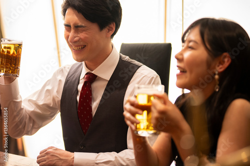 Obraz na płótnie A man and woman sit side by side in a stylish bar, laughing and enjoying a drink together. Both are holding mugs of beer, and the mood is lighthearted and full of camaraderie, suggesting a casual date w salonie