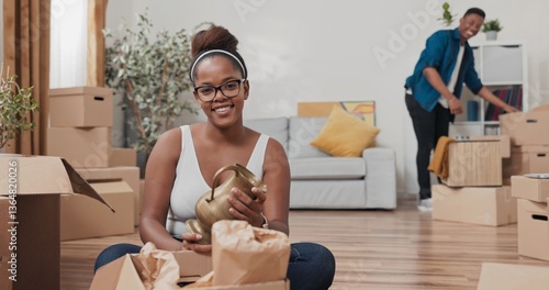 A wife sits on the floor of her freshly bought apartment, takes out a decorative vase from paper, and smiles gently. In the background, her husband continues cleaning the living room.
