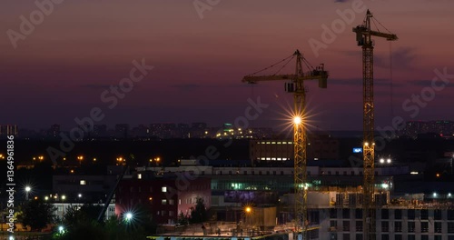 Night time lapse. Two large cranes work at night on the construction of a multi-story office building. A lot of workers are running around on the upper level of the construction site. Beautiful sunset