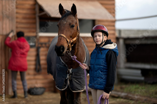 Brown horse and young girl with blanket near wooden stable. Child holds purple lead rope and gently pets horse in riding helmet and blue vest standing. Rural, equestrian sport, outdoor activity.