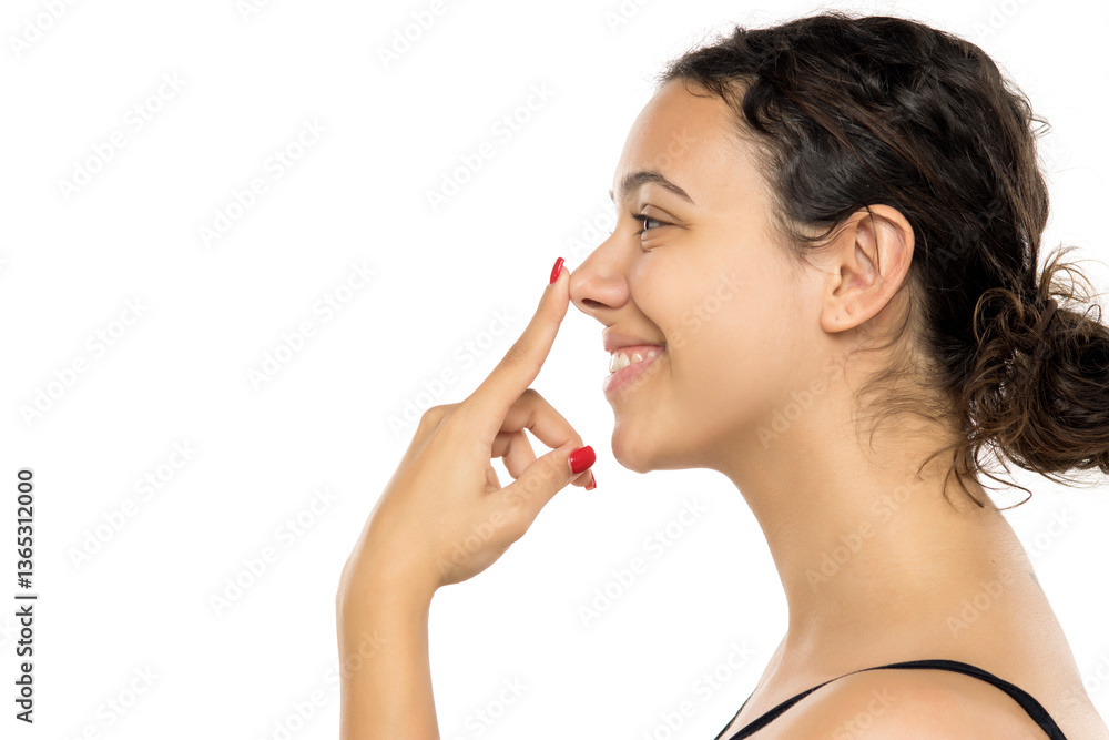 Profile view of young woman smiling while touching nose, eyes looking away. Dark curly hair in bun. Positive expression suggesting rhinoplasty consideration. Studio portrait. Profile view of young woman smiling while touching nose, eyes looking away. Dark curly hair in bun. Positive expression suggesting rhinoplasty consideration. Studio portrait.