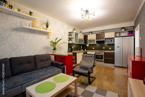 Apartment interior. Living room combined with kitchen. Furniture and kitchen appliances: dark gray sofa, armchair, coffee table, red chests of drawers, chandelier, kitchen set and refrigerator.