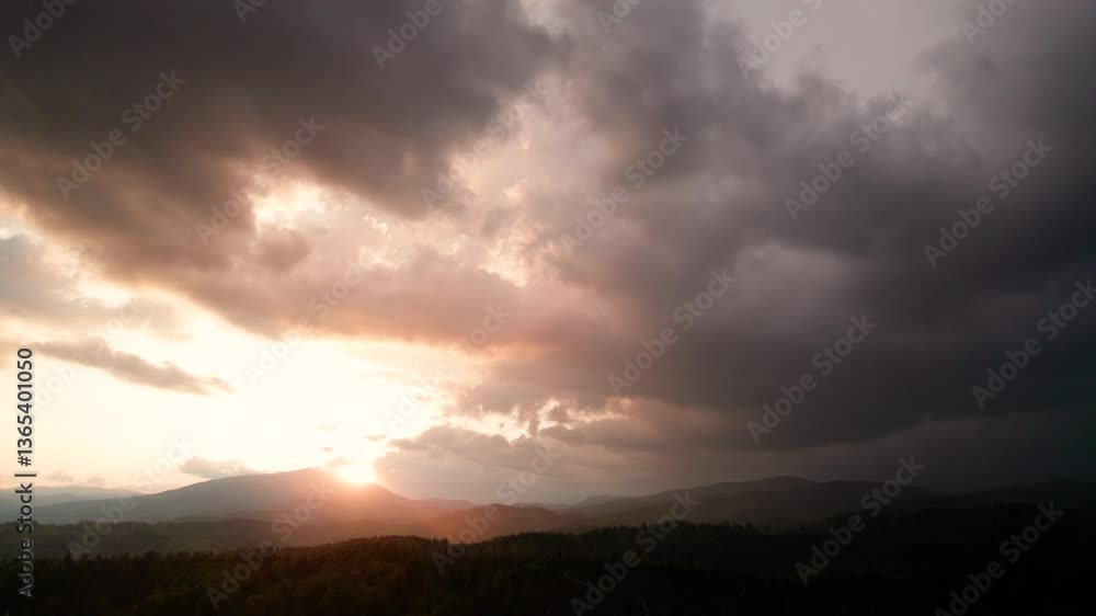 Aerial view of dramatic sky with thick, dark clouds looms over mountainous landscape. Sun peeks through, casting warm, golden light on rolling hills and dense forests below, creating striking contrast Aerial view of dramatic sky with thick, dark clouds looms over mountainous landscape. Sun peeks through, casting warm, golden light on rolling hills and dense forests below, creating striking contrast
