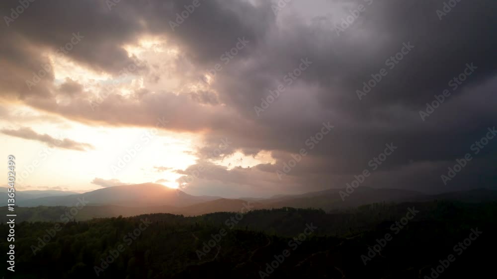 Aerial view of dramatic sky with thick, dark clouds looms over mountainous landscape. Sun peeks through, casting warm, golden light on rolling hills and dense forests below, creating striking contrast Aerial view of dramatic sky with thick, dark clouds looms over mountainous landscape. Sun peeks through, casting warm, golden light on rolling hills and dense forests below, creating striking contrast