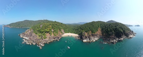 A stunning aerial top view of Butterfly Beach in Goa, showcasing golden sands waters and lush green hills in the backdrop. People are seen enjoying the beach while a boat sails nearby.
