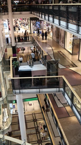 Vertical screen: Multi-level shopping mall with escalators, fashion stores, and walking visitors. The clean architecture and busy flow of people reflect the vibrant retail environment.

