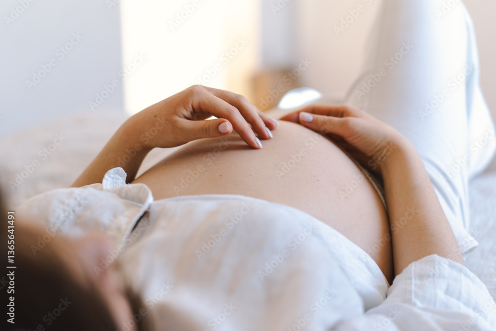 Obraz premium A pregnant woman sits comfortably on a bed, wearing relaxed white clothing. She gently holds her belly, embodying tranquility and connection with the child