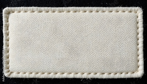 A close-up of an off-white fabric label that is blank and has a stitched border, set against a black backdrop