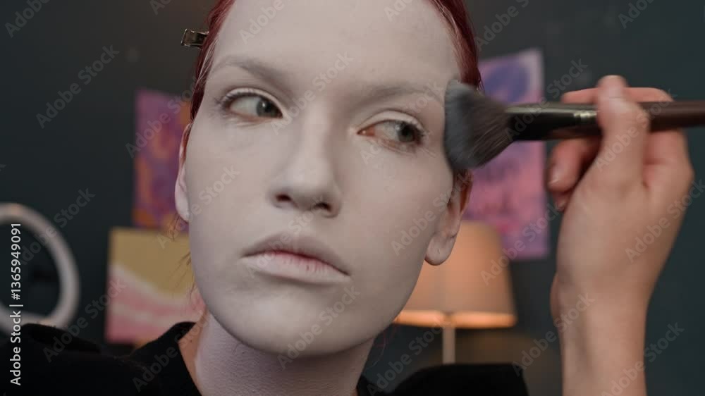 Close-up tilt up view of red-haired gen Z girl wearing black jumper blending white paint with makeup brush after putting it on her face, ears and neck Close-up tilt up view of red-haired gen Z girl wearing black jumper blending white paint with makeup brush after putting it on her face, ears and neck