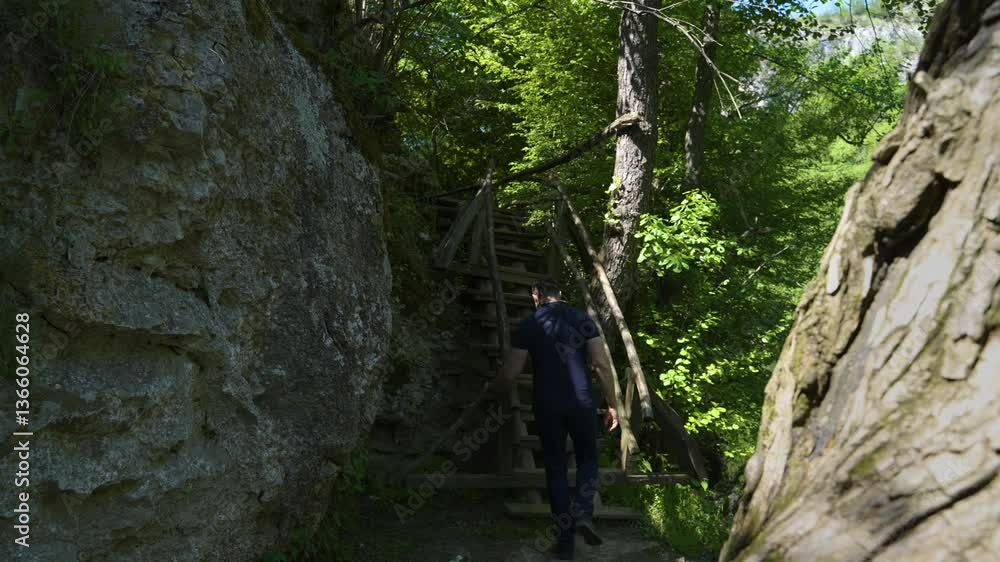 A man walks along the Geopark Iskar-Panega eco-trail, surrounded by lush greenery and rugged rock formations. A rustic wooden staircase leads further into the scenic natural landscape. A man walks along the Geopark Iskar-Panega eco-trail, surrounded by lush greenery and rugged rock formations. A rustic wooden staircase leads further into the scenic natural landscape.
