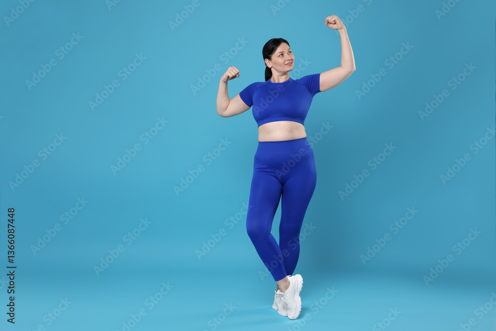 Fototapeta premium Plus size woman in gym clothes showing her biceps on light blue background, space for text