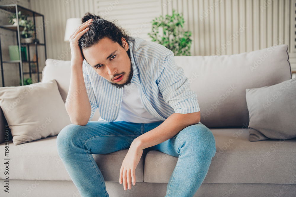 Young man casually dressed sitting on a sofa in a bright modern living room, looking thoughtful, relaxed and deep in contemplation Young man casually dressed sitting on a sofa in a bright modern living room, looking thoughtful, relaxed and deep in contemplation