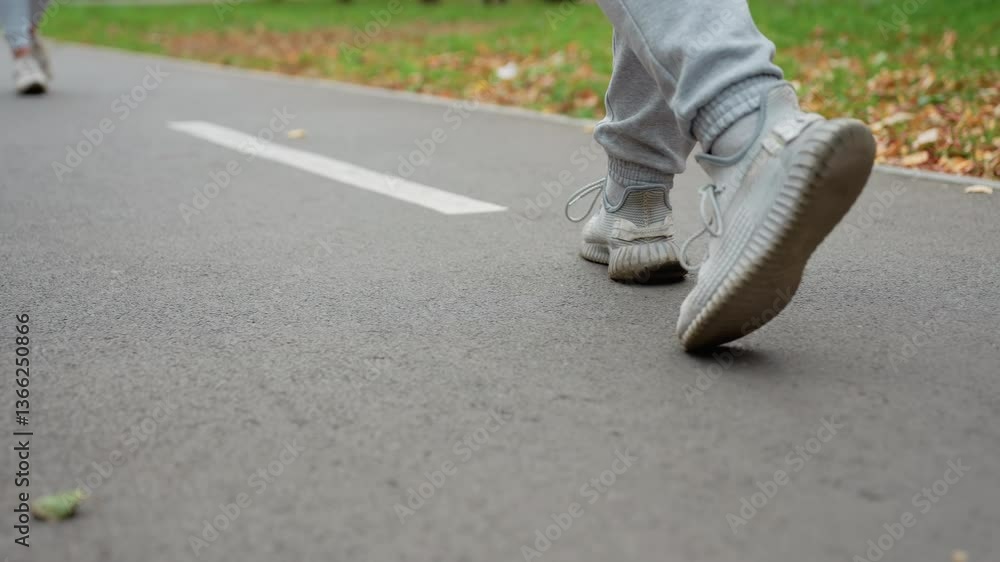 Side view of person walking on road with white markings wearing sportswear and sneakers, surrounded by greenery and scattered autumn leaves in peaceful park setting Side view of person walking on road with white markings wearing sportswear and sneakers, surrounded by greenery and scattered autumn leaves in peaceful park setting