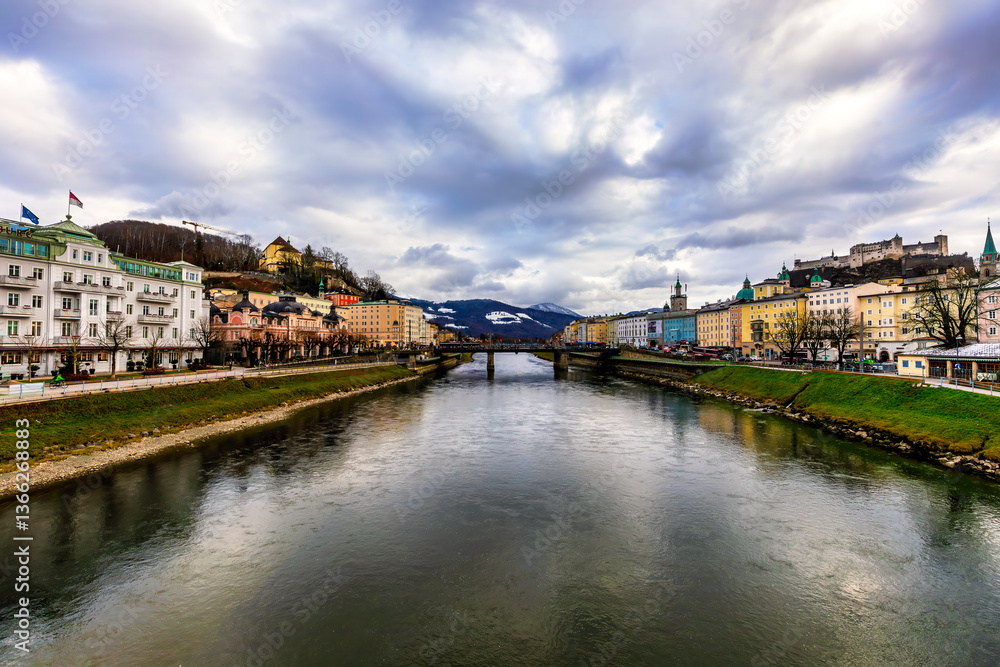 Fototapeta premium A stunning view of Salzburg’s Salzach River, with St. Markus Church on a hill to the left and the Evangelische Christuskirche’s spire on the right. A bridge links both banks.