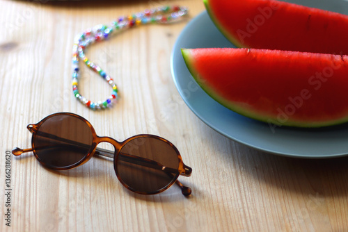 Plate with watermelon slices, glass of orange juice or cocktail, straw hat, colorful beaded necklace and sunglasses on the wooden table. Summer essentials concept. Selective focus.
