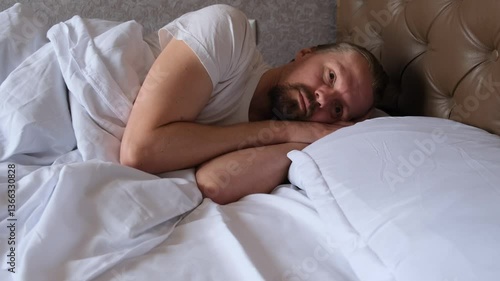A young man lying in bed opens his eyes waking up and then closes his eyes again continuing to sleep. Tense and restless sleep. Insomnia