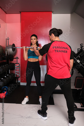 Personal trainer assisting young woman lifting weights during fitness training session in modern gym