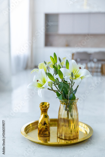 Bouquet of white artificial lilies in a golden vase on a table on a light background.
