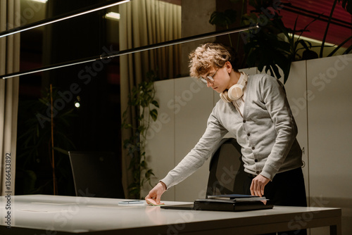 Canvas Print A young White man with glasses and headphones around his neck organizes sticky notes at a sleek white desk in a modern office at night