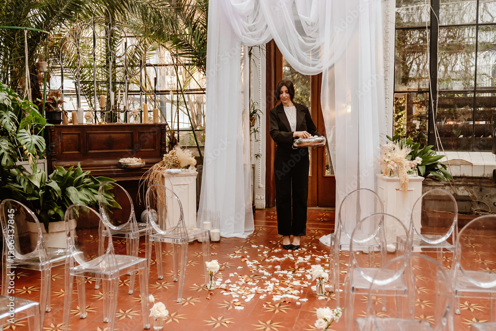 Obraz premium A woman in a black suit sets up the wedding ceremony with flower petals on the floor, preparing for the upcoming celebration in a beautiful garden-like venue with plants and a piano.