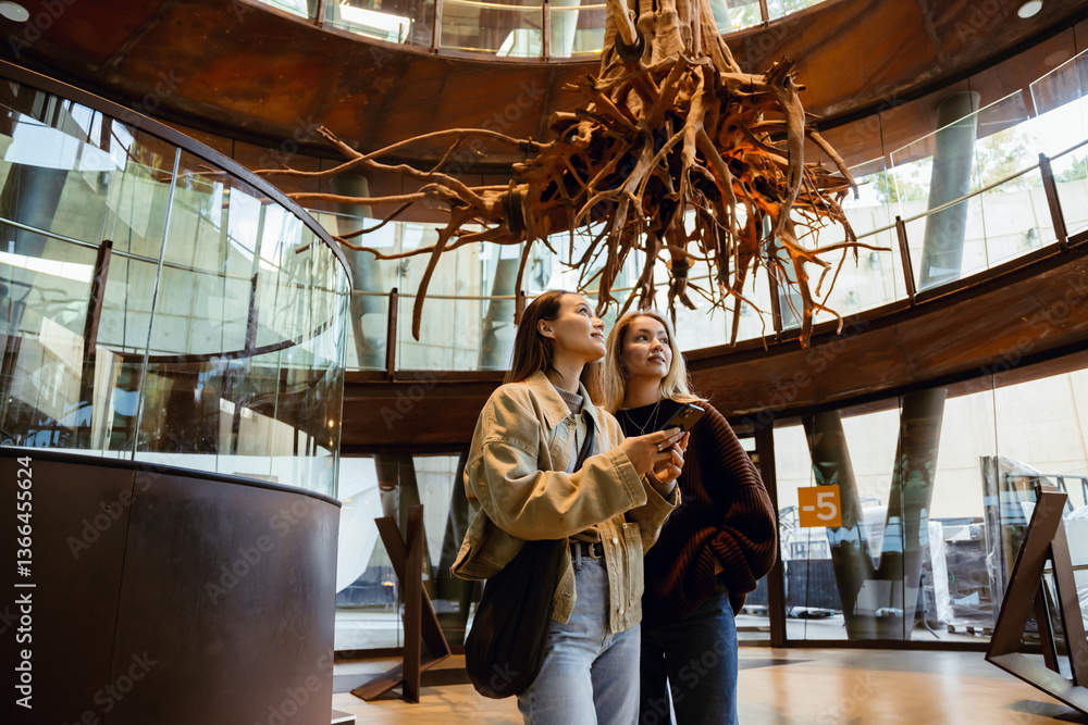 A woman stands and holds a phone while her female friend stands next to her with her hand in her pocket, they are smiling and looking away, while traveling A woman stands and holds a phone while her female friend stands next to her with her hand in her pocket, they are smiling and looking away, while traveling