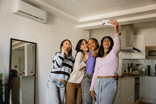 A group of four female friends stand and pose while smiling and looking at a phone held by one of them, near the kitchen, at a girls' party