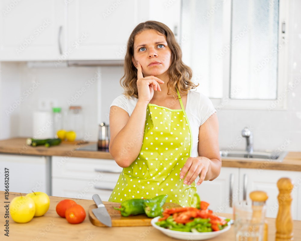 Portrait of young pensive woman standing near kitchen countertop while preparing vegetables for healthy dinner at home Portrait of young pensive woman standing near kitchen countertop while preparing vegetables for healthy dinner at home