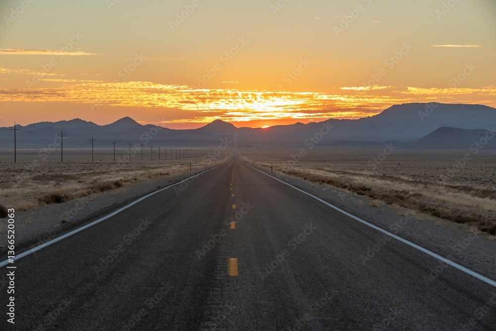 A long, straight highway stretches into the distance in Tonopah, Nevada, USA, leading towards mountains as the sun sets. The golden sky contrasts with the dark landscape. A long, straight highway stretches into the distance in Tonopah, Nevada, USA, leading towards mountains as the sun sets. The golden sky contrasts with the dark landscape.