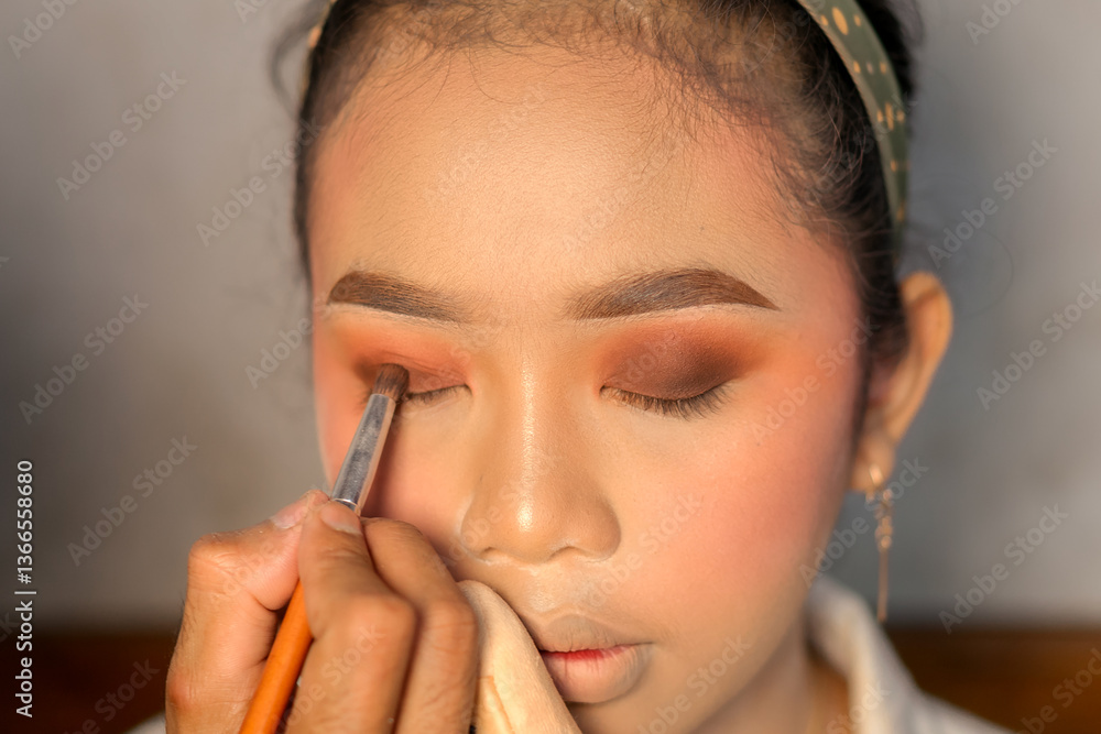 Obraz premium A close-up of a makeup artist applying dark eyeshadow to a woman’s eyelid with a brush. The woman wears a green polka dot headband and gold earrings.