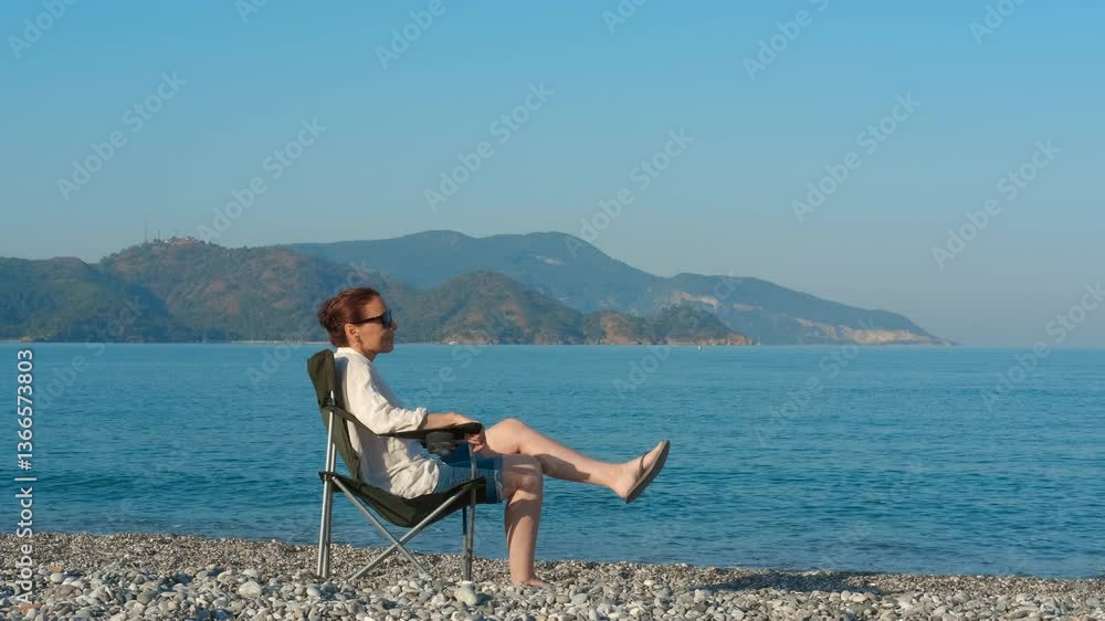 Woman folds her beach chair after enjoying a relaxing day at a pebble beach. Leaving the seaside, she takes in the beautiful view of the ocean and the clear blue sky Woman folds her beach chair after enjoying a relaxing day at a pebble beach. Leaving the seaside, she takes in the beautiful view of the ocean and the clear blue sky