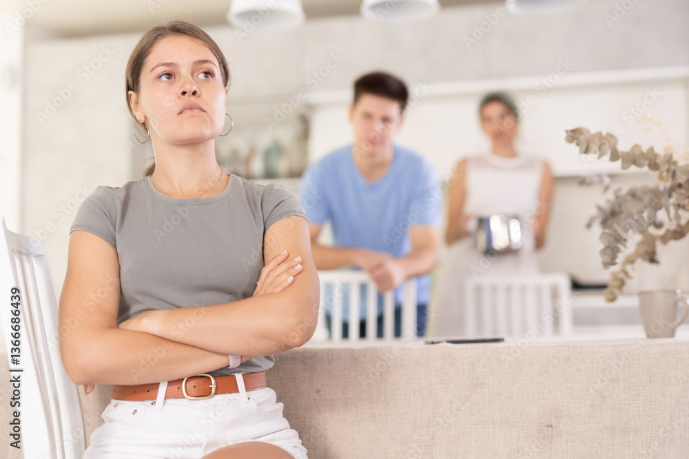 Obraz premium Sad offended young woman sitting at kitchen table during argument with dissatisfied husband and mother-in-law standing behind on blurred background. Concept of family fights