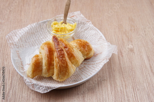 A golden brown croissant served on a wooden board with a small glass bowl of butter and set on a rustic wooden table.