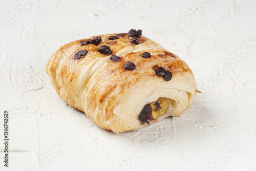 Close-up of a chocolate chip pastry with a visible banana filling, placed on a white textured surface. The pastry has a golden, flaky crust topped with chocolate chips.