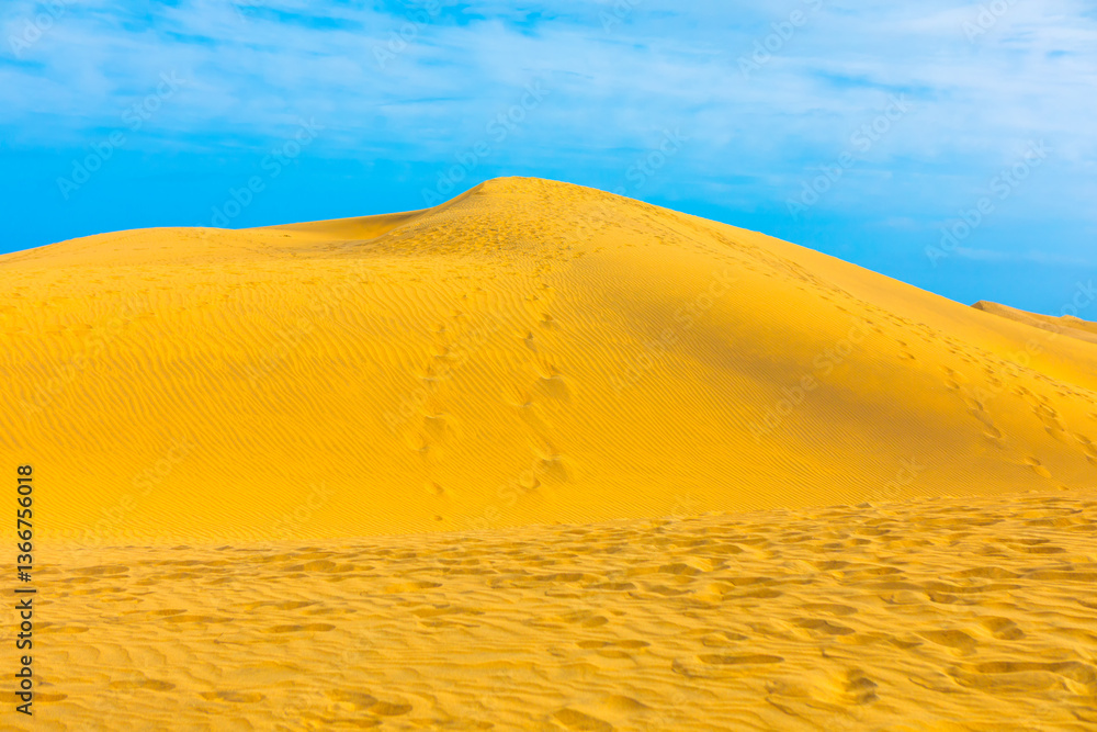 Stunning yellow sand dune under a vibrant blue sky dotted with white, scattered clouds. Human footprints trailing across the dune bring a sense of adventure to this serene, natural landscape