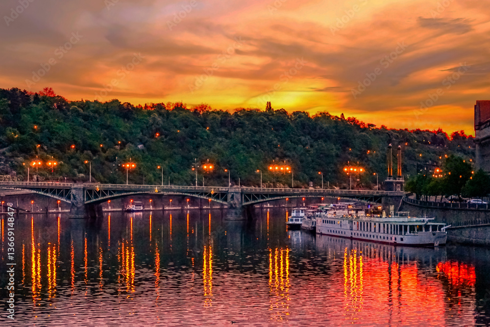 Fototapeta premium The elegant Svatopluk Čech Bridge gracefully spans the Vltava River in Prague, Czech Republic, at twilight, with its lights mirroring on the water alongside docked tourist boats.