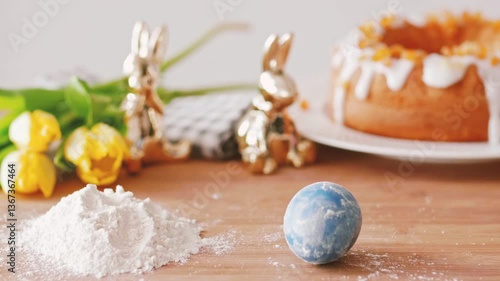 A festive Easter-themed still life featuring a blue marbled egg on a wooden surface, with flour, golden bunny figurines, yellow tulips, and a glazed cake in the softly blurred background.
