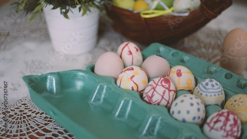 A pastel-colored carton of hand-painted Easter eggs with abstract patterns in red, blue, and yellow, placed on a lace-covered table with a softly blurred background.