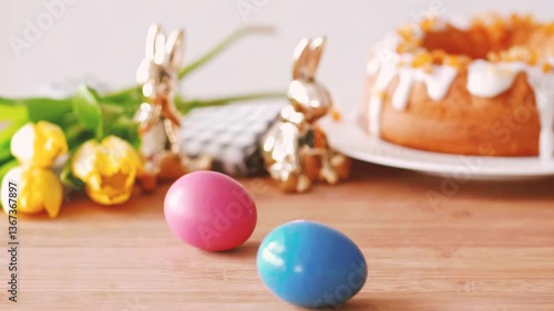 A festive Easter scene featuring two colorful dyed eggs on a wooden surface, with golden bunny figurines, fresh yellow tulips, and a glazed cake in the softly blurred background.