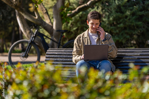 Chico con ordenador negro hablando con telefono movil en un banco en el parque un dia soleado con una bici electrica de fondo