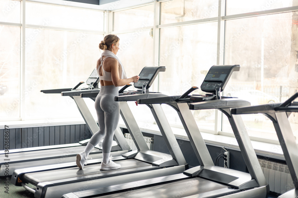 Fototapeta premium Happy European woman in sportswear and wireless headphones and towel around neck doing hard cardio workout on treadmill, lady working out in empty gym
