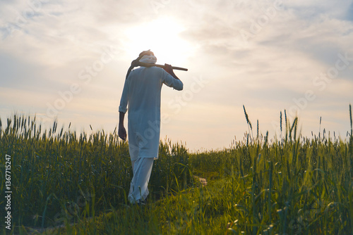 A traditional Pakistani farmer walks through a wheat field at sunset, carrying a farming tool on his shoulder.