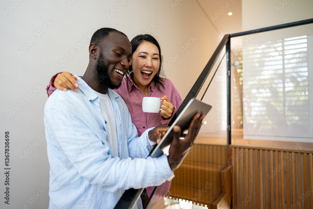 Multinational couple enjoying their morning routine, smiling and using laptop and tablet at home