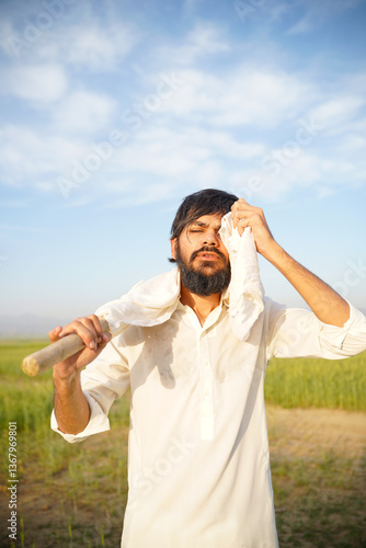 A hardworking Pakistani Indian farmer wipes sweat from his face while carrying a hoe, standing in a sunlit agricultural field.