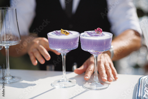 A bartender is meticulously preparing two vibrant purple cocktails on the table