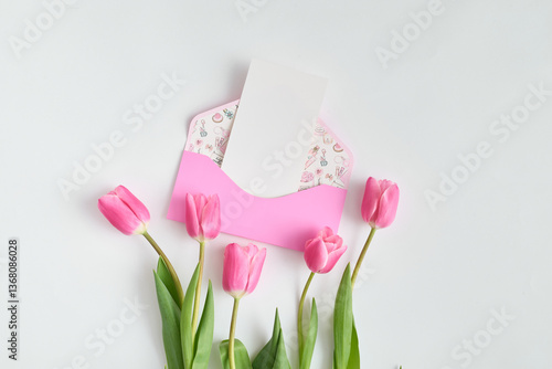 Pink tulips are lying on a white table next to an envelope containing a greeting card. Minimal composition layout
