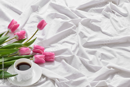 A joyful Mother's Day morning features a cup of coffee accompanied by fresh pink tulips resting on a cozy white bedspread.