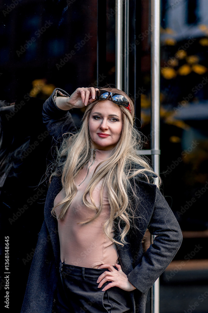 Portrait of blonde stylish woman posing near glass doors wearing black pants and coat