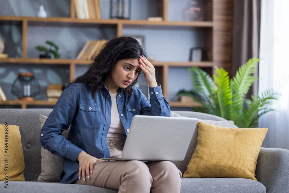 Upset young Indian woman sitting on the couch at home, holding a laptop on her lap and looking worriedly at the screen while holding her head in her hand Upset young Indian woman sitting on the couch at home, holding a laptop on her lap and looking worriedly at the screen while holding her head in her hand